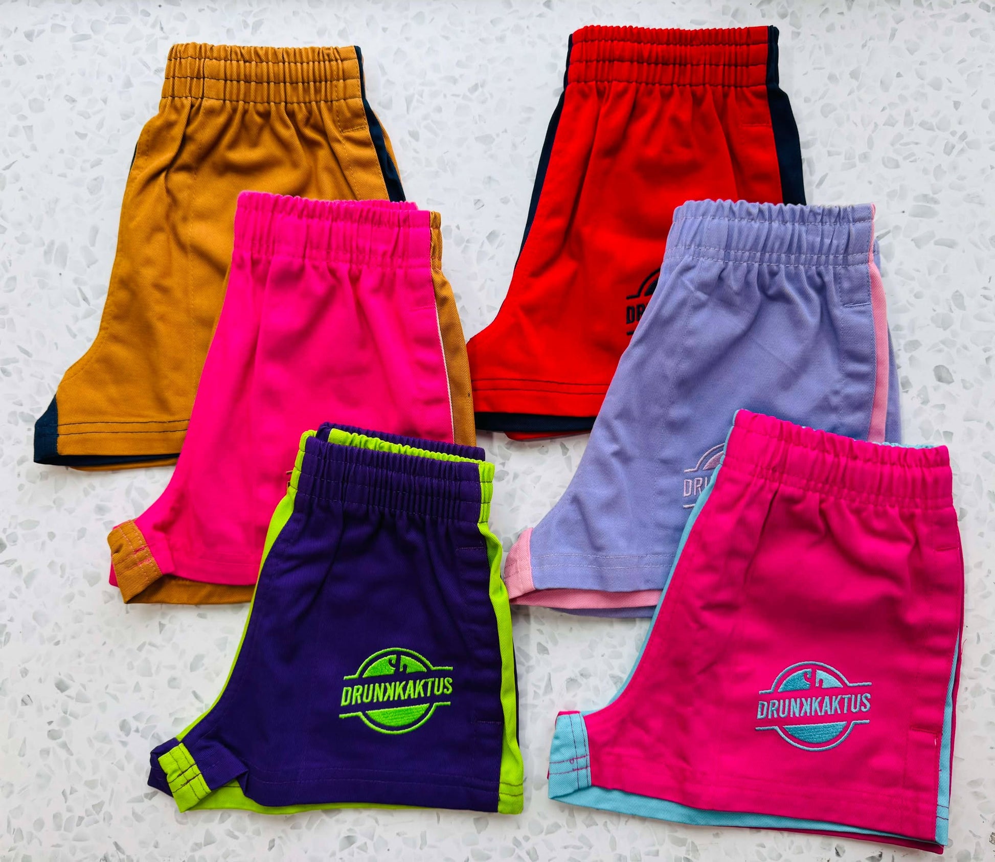 Fan layout of kids mustard pink ruggers cotton drill shorts displayed alongside kids green purple ruggers, kids ruggers blue pink cotton drill shorts, navy mustard cotton shorts, and kids pink purple shorts.